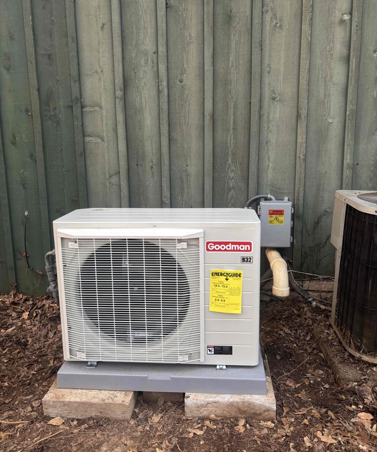 Front view of new Goodman R32 condenser with EnergyGuide sticker showing 19.0 SEER2 rating, installed on proper composite pad next to the old unit still visible for comparison
