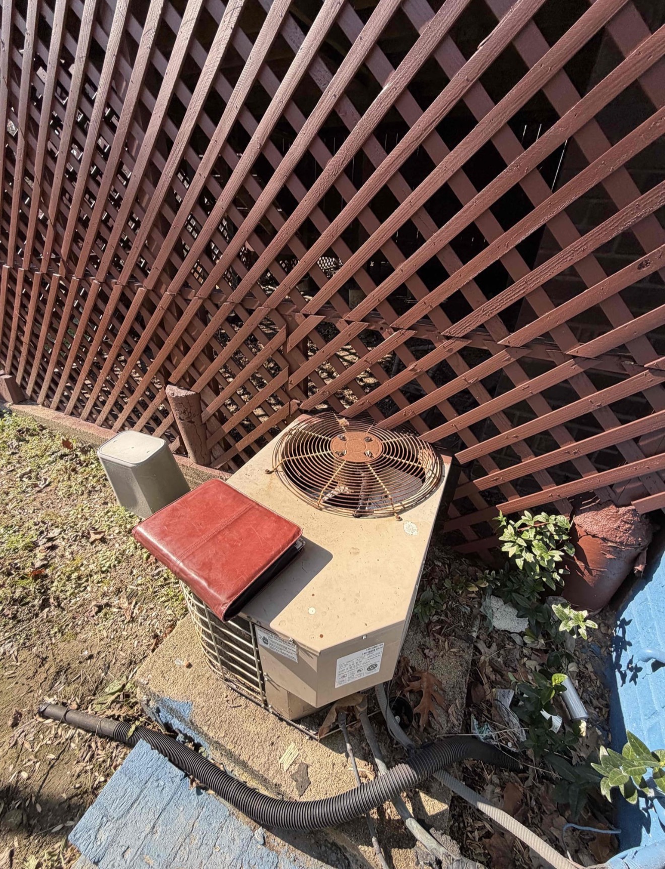 Old weathered condenser unit crammed under lattice screening with zero clearance and debris piled around it, sitting on bare concrete with no proper equipment pad